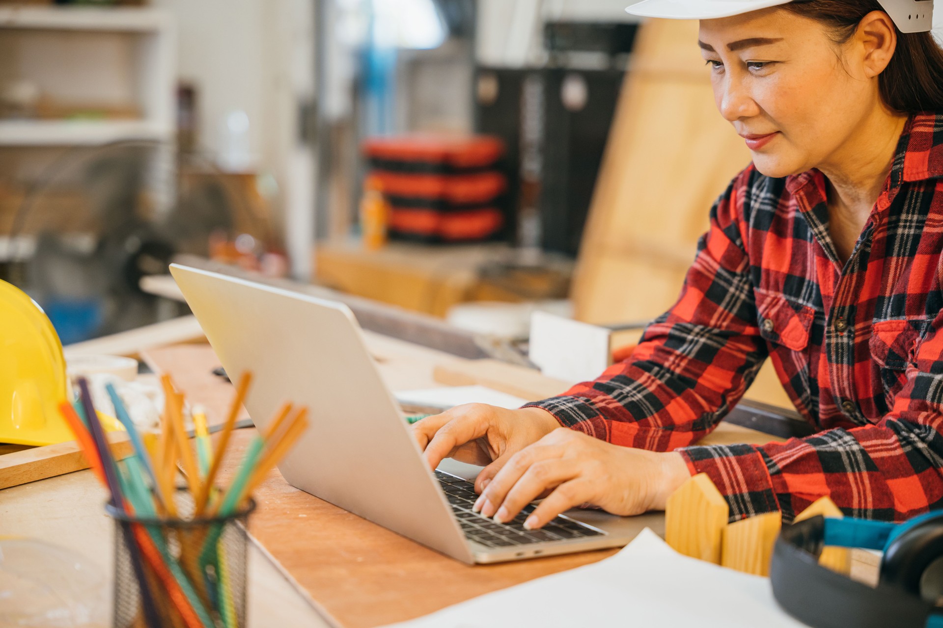 Carpenter woman in a white hardhat and red plaid shirt uses a laptop in a woodshop, surrounded by tools, wood scraps, and pencils. Ideal for online learning